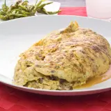 Tortilla de bacalao vasca, dorada y jugosa, servida en plato blanco sobre mantel rojo. Acompañada de pimientos verdes fritos.