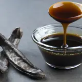 Miel de caña oscura y espesa cayendo de una cuchara de madera a un cuenco de cristal, junto a vainas de algarroba. Descubre sus propiedades.