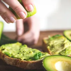 Una mano exprime lima sobre tostadas con aguacate machacado. El aguacate tiene fibra, ideal para un desayuno saludable.