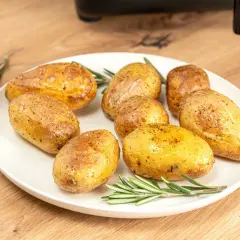 Patatas asadas en freidora de aire, doradas y crujientes, servidas con romero fresco en un plato blanco sobre una mesa de madera.