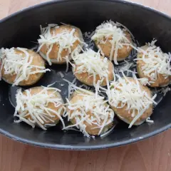 Patatas rellenas de atún listas para hornear, cubiertas con queso rallado en una bandeja oscura.