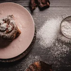 Un rollo de canela espolvoreado con azúcar glas, que es fecula de maiz, sobre un plato rosa. Un colador con más polvo blanco está cerca.