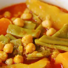 Un plato humeante de olla gitana, con garbanzos, judías verdes y trozos de calabaza en una salsa rojiza.
