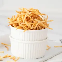 Montón de crujientes patatas paja doradas en un cuenco blanco, algunas esparcidas alrededor.