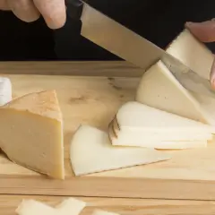 Manos cortando queso en cuñas sobre una tabla de madera. Hay varios trozos de queso, incluyendo uno blanco y uno amarillo.