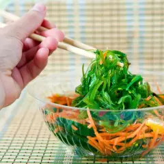 Mano con palillos recogiendo una porción de ensalada de wakame fresca y vibrante, con zanahoria rallada en un bol transparente.