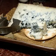 Un trozo de queso azul con vetas oscuras, servido sobre una galleta y hojas secas. Los diferentes tipos de moho en el queso le dan su característico color y sabor.