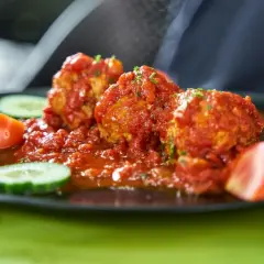 Albondigas con tomate, jugosas y cubiertas de salsa roja, acompañadas de rodajas de pepino y tomate fresco en un plato negro.