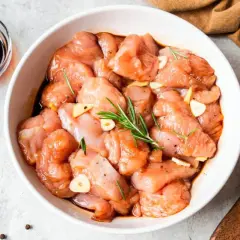 Pechuga de pollo cruda marinando en un tazón blanco con ajo, romero y salsa de soja.