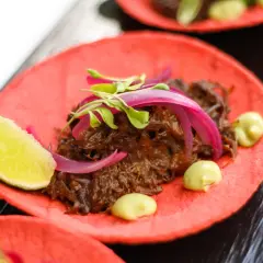 Deliciosos tacos de rabo de toro sobre tortilla roja, con cebolla morada, brotes verdes y un toque de aguacate.