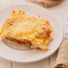 Porción de pastel de berenjenas gratinado con queso, capas de jamón y berenjena, servido en plato blanco.