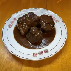 Tres trozos de rabo de toro estofado en una salsa oscura y rica, servidos en un plato blanco con borde dorado y decorado con rosas.