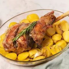 Pierna de cordero al horno dorada, servida con patatas asadas, romero y ajo en una fuente de cristal.