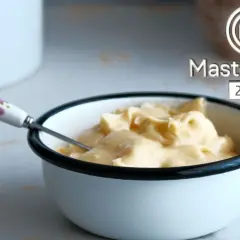 Crema sedosa lista para emulsionar en cocina. Un tazón blanco con borde negro contiene una suave crema amarilla, con una cuchara de mango floral.