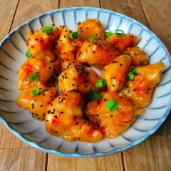 Bocados de pollo sweet chilli glaseados, espolvoreados con semillas de sésamo negro y cebolleta picada, servidos en un plato decorado.