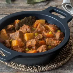 Un reconfortante estofado de ternera con patatas, guisantes y laurel en una cazuela negra sobre madera rústica.