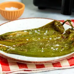 Dos pimientos verdes fritos, salpicados de sal gruesa, reposan en un plato blanco con borde marrón. Un paño a cuadros rojos y blancos y un cuenco de sal completan la escena.