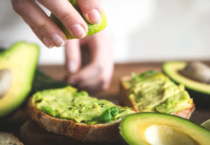 Una mano exprime lima sobre tostadas con aguacate machacado. El aguacate tiene fibra, ideal para un desayuno saludable.