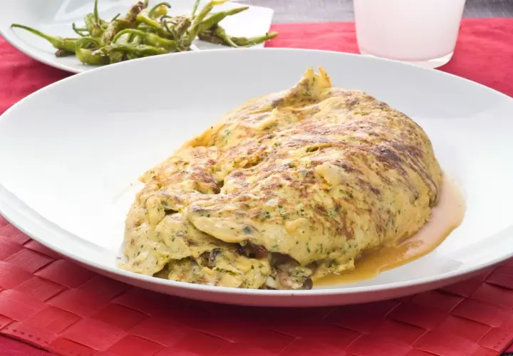 Tortilla de bacalao vasca, dorada y jugosa, servida en plato blanco sobre mantel rojo. Acompañada de pimientos verdes fritos.