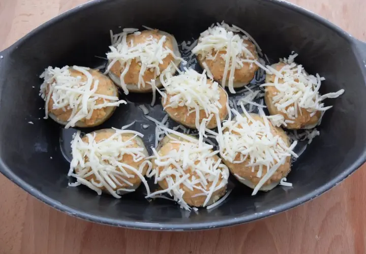 Patatas rellenas de atún listas para hornear, cubiertas con queso rallado en una bandeja oscura.