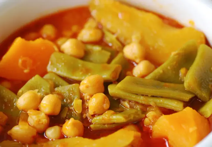 Un plato humeante de olla gitana, con garbanzos, judías verdes y trozos de calabaza en una salsa rojiza.