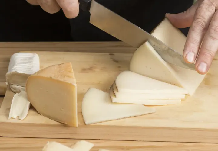 Manos cortando queso en cuñas sobre una tabla de madera. Hay varios trozos de queso, incluyendo uno blanco y uno amarillo.