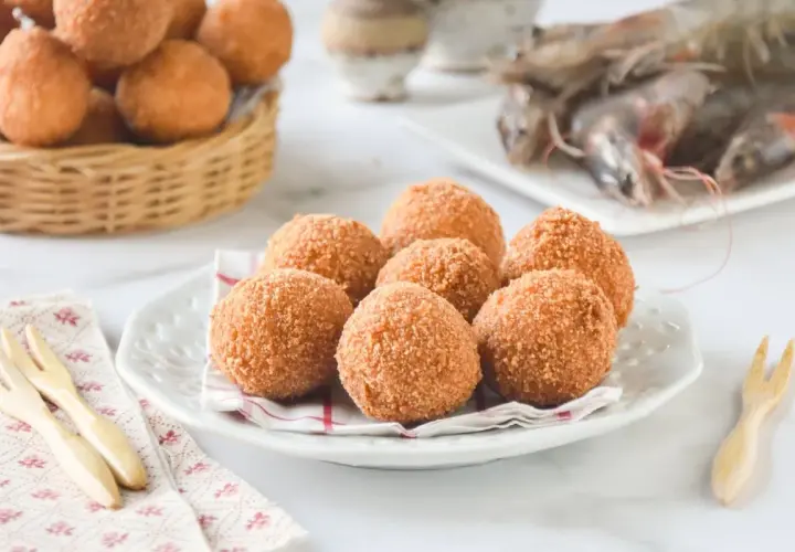 Croquetas de gambas doradas y crujientes en un plato blanco, con más en una cesta de mimbre al fondo.