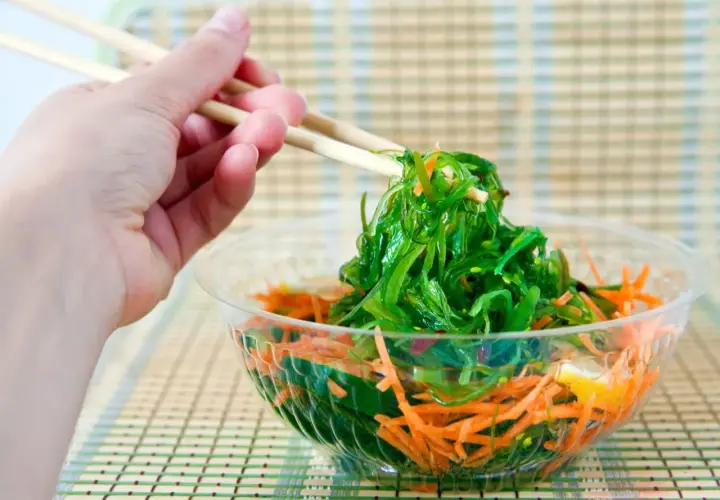 Mano con palillos recogiendo una porción de ensalada de wakame fresca y vibrante, con zanahoria rallada en un bol transparente.