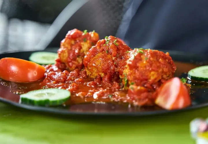 Albondigas con tomate, jugosas y cubiertas de salsa roja, acompañadas de rodajas de pepino y tomate fresco en un plato negro.