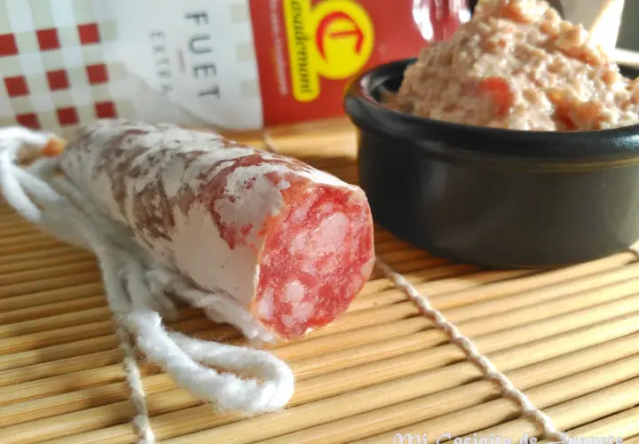 Un trozo de fuet extra con su interior rosado y veteado, junto a un cuenco de paté.