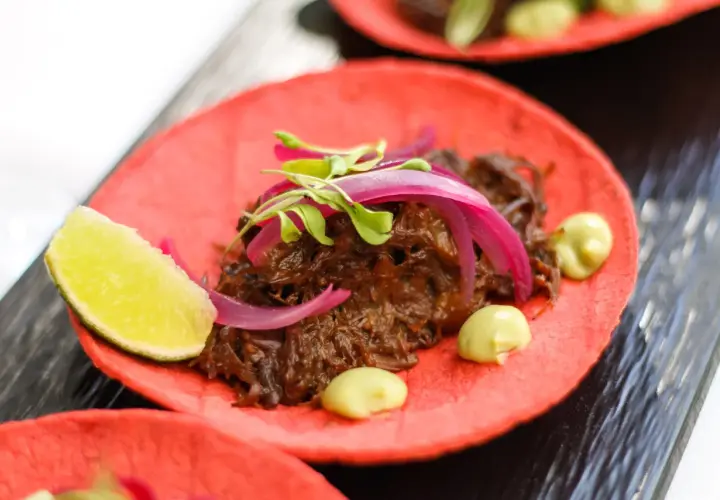 Deliciosos tacos de rabo de toro sobre tortilla roja, con cebolla morada, brotes verdes y un toque de aguacate.
