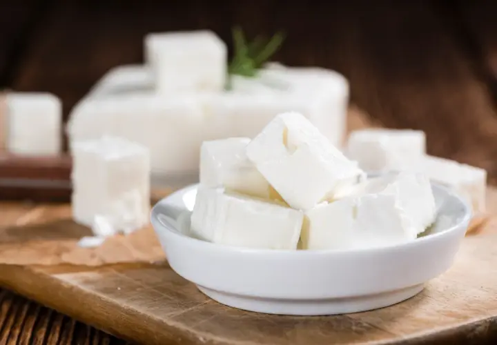 Cubos de queso blanco en un cuenco, con un bloque de queso de leche cruda al fondo. El queso de leche cruda puede presentar peligros.