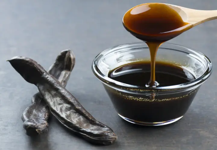 Miel de caña oscura y espesa cayendo de una cuchara de madera a un cuenco de cristal, junto a vainas de algarroba. Descubre sus propiedades.