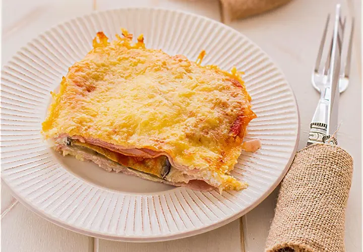 Porción de pastel de berenjenas gratinado con queso, capas de jamón y berenjena, servido en plato blanco.