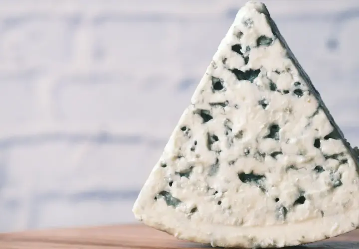 Un trozo triangular de queso azul, con sus características vetas de moho azul verdoso, descansa sobre una superficie de madera.