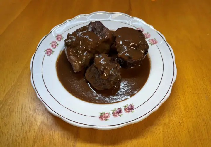 Tres trozos de rabo de toro estofado en una salsa oscura y rica, servidos en un plato blanco con borde dorado y decorado con rosas.