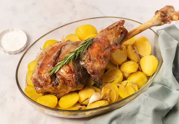 Pierna de cordero al horno dorada, servida con patatas asadas, romero y ajo en una fuente de cristal.