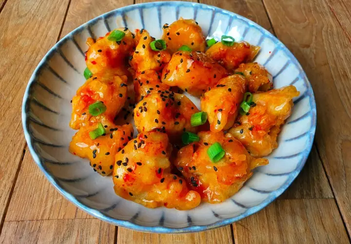 Bocados de pollo sweet chilli glaseados, espolvoreados con semillas de sésamo negro y cebolleta picada, servidos en un plato decorado.