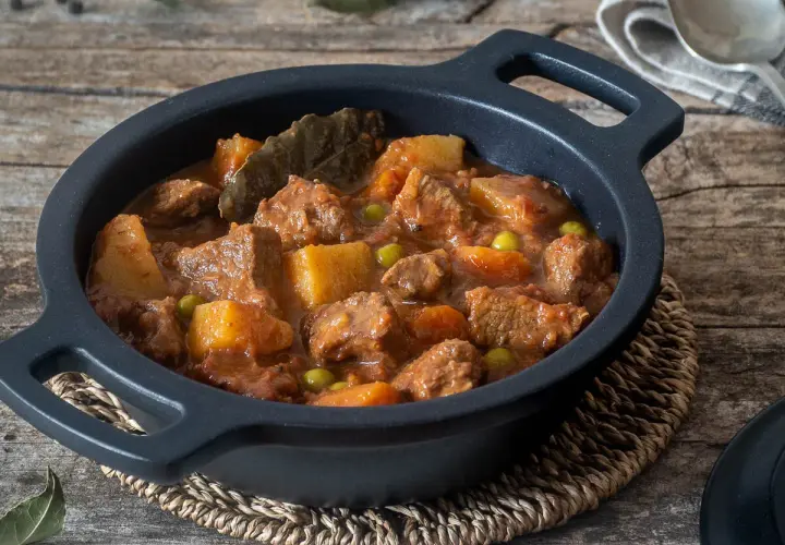 Un reconfortante estofado de ternera con patatas, guisantes y laurel en una cazuela negra sobre madera rústica.
