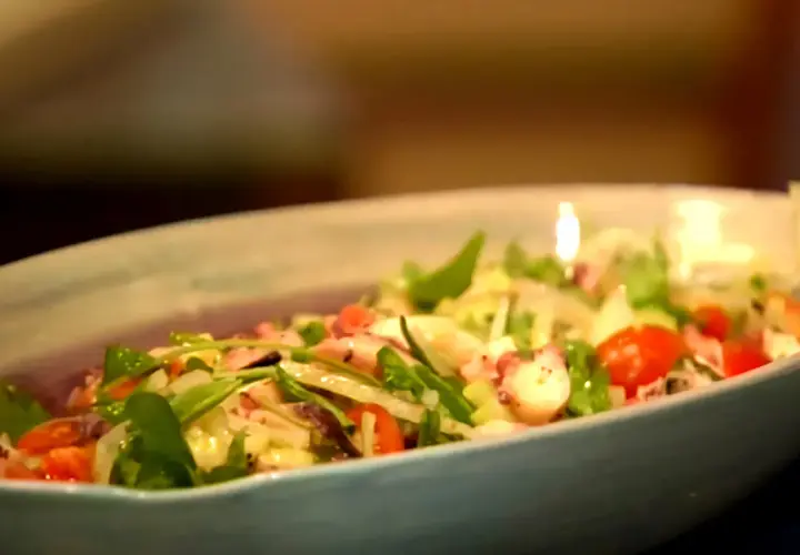 Deliciosa insalata di mare con tentáculos de pulpo, tomates cherry y hojas verdes frescas en un plato azul claro.