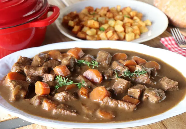 Ternera estofada en salsa con zanahorias y patatas fritas al fondo.