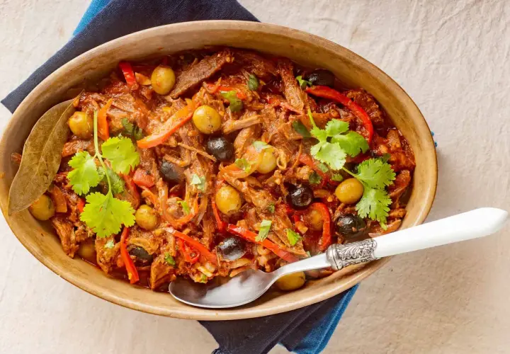 Delicioso plato de ropa vieja con aceitunas verdes y negras, pimientos rojos y cilantro fresco, servido en una cazuela de barro.