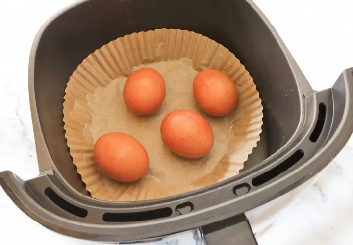 Cuatro huevos listos para convertirse en huevo duro airfryer, colocados sobre papel de hornear en la cesta de la freidora de aire.