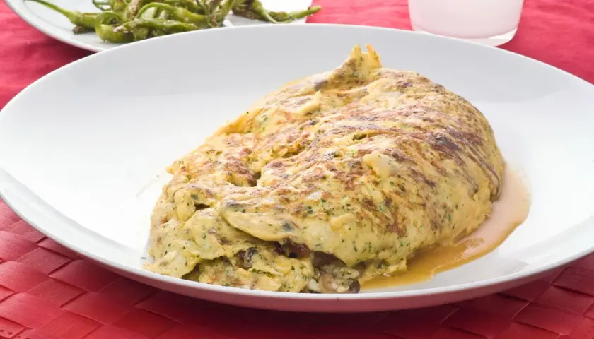 Tortilla de bacalao vasca, dorada y jugosa, servida en plato blanco sobre mantel rojo. Acompañada de pimientos verdes fritos.