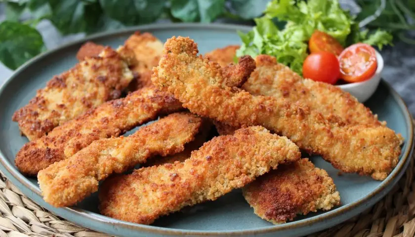 Crujientes fingers de pollo dorados, servidos en un plato azul con una guarnición de lechuga y tomates cherry. ¡Perfectos para cualquier ocasión!
