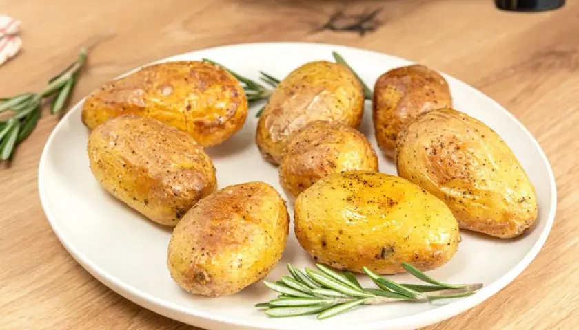 Patatas asadas en freidora de aire, doradas y crujientes, servidas con romero fresco en un plato blanco sobre una mesa de madera.
