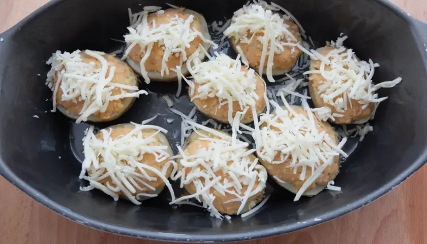 Patatas rellenas de atún listas para hornear, cubiertas con queso rallado en una bandeja oscura.
