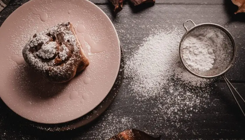 Un rollo de canela espolvoreado con azúcar glas, que es fecula de maiz, sobre un plato rosa. Un colador con más polvo blanco está cerca.