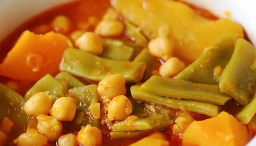 Un plato humeante de olla gitana, con garbanzos, judías verdes y trozos de calabaza en una salsa rojiza.