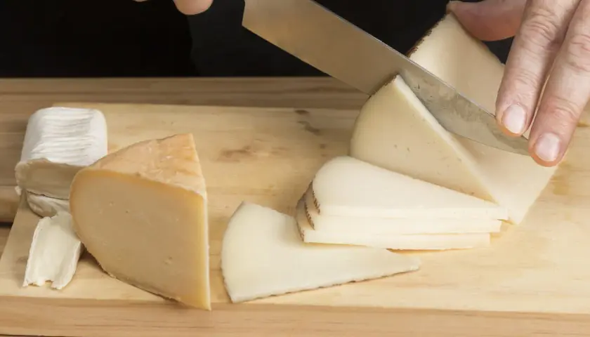Manos cortando queso en cuñas sobre una tabla de madera. Hay varios trozos de queso, incluyendo uno blanco y uno amarillo.